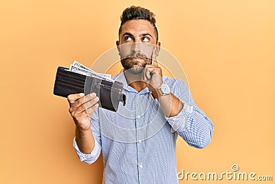 Handsome man with beard holding wallet with dollars serious face thinking about question with hand on chin, thoughtful about Stock Photo