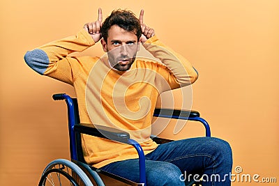 Handsome man with beard sitting on wheelchair doing funny gesture with finger over head as bull horns Stock Photo