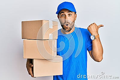 Handsome man with beard wearing courier uniform holding delivery packages surprised pointing with hand finger to the side, open Stock Photo