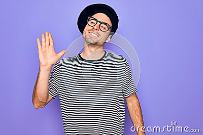 Handsome man with blue eyes wearing striped t-shirt and french beret over purple background Waiving saying hello happy and Stock Photo