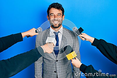 Handsome man business with beard being interviewed by reporters holding microphone looking positive and happy standing and smiling Stock Photo