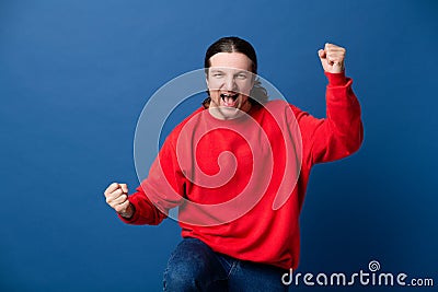 Handsome man excited doing winner gesture with arms, wearing casual red sweater very happy standing over isolated blue background, Stock Photo