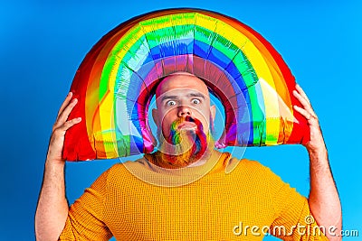 handsome man with raindow colors in beard and yellow sweater holding balloon inflatable above of his head in blue studio Stock Photo