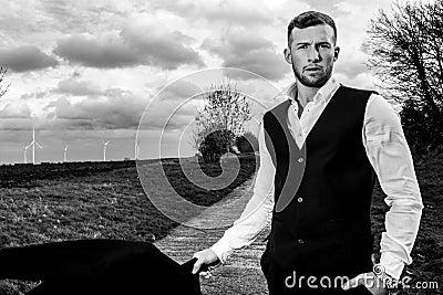 Handsome man in suit and waistcoat standing with fields and stormy sky behind him looking at camera. Stock Photo