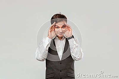 Handsome man in a white shirt and grey vest hiding behind his hands isolated over grey background. Stock Photo