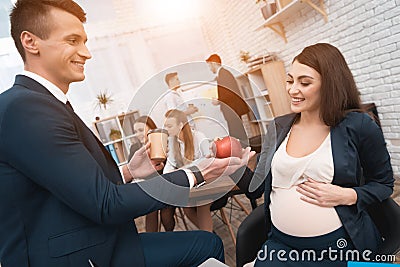 Handsome man in suit gives apple to young pregnant woman in office. Pregnancy at work. Stock Photo