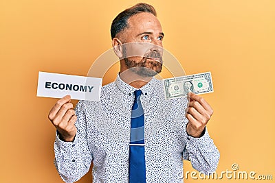 Handsome middle age man holding economy message and holding 1 dollar smiling looking to the side and staring away thinking Stock Photo