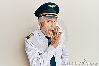 Handsome middle age mature man wearing airplane pilot uniform hand on mouth telling secret rumor, whispering malicious talk Stock Photo