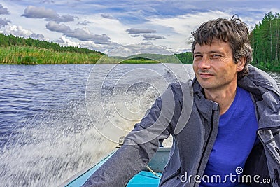 Handsome middle-aged man sitting at boat stern and floating along northern river on beautiful landscape background in summer day. Stock Photo