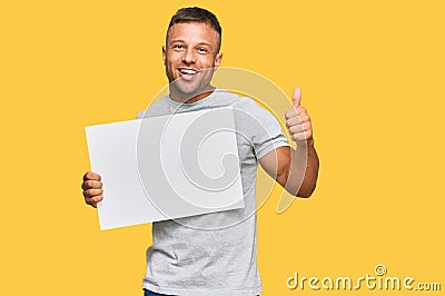 Handsome muscle man holding blank empty banner smiling happy and positive, thumb up doing excellent and approval sign Stock Photo