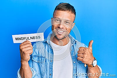 Handsome muscle man holding mindfulness word on paper smiling happy and positive, thumb up doing excellent and approval sign Stock Photo