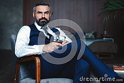Handsome pensive man is touching his beard, looking away and thinking while sitting in armchair indoors. Stock Photo