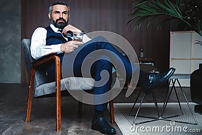 Handsome pensive man is touching his beard, looking away and thinking while sitting in armchair indoors. Stock Photo