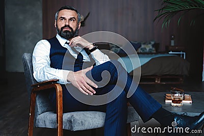 Handsome pensive man is touching his beard, looking away and thinking while sitting in armchair indoors. Stock Photo