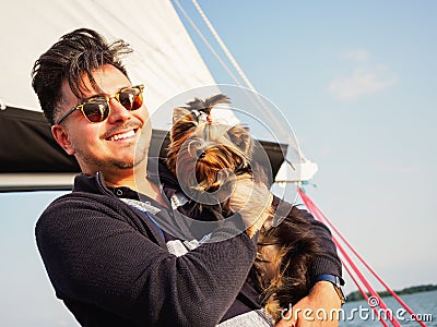 Handsome person hugs his small dog yorkshire terrier on a sailing yacht during vacations, travel with pets concept Stock Photo