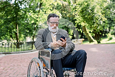 Handsome senior concentrated bearded man sitting in wheelchair in park, bowing his head while looking at screen of his Stock Photo