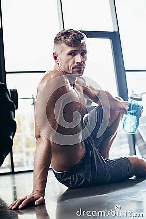 handsome shirtless sportsman sitting on floor in gym and holding sport bottle, looking Stock Photo