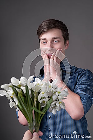 Handsome shocked young man with dark hair in blue shirt holding cheeks and looking at presenting bouguet of flowers Stock Photo