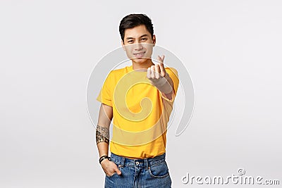 Handsome and stylish hipster asian guy in yellow t-shirt, with tattoos, extend arm and showing korean heart love sign Stock Photo