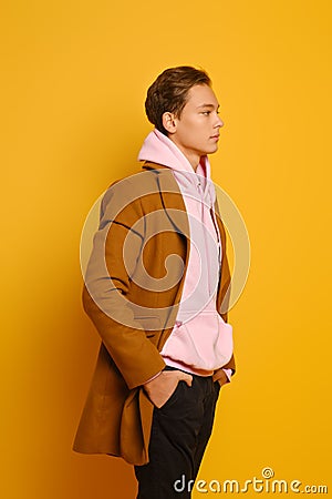 Handsome stylish man in brown trenchcoat and pink sweatshirt with hood standing in profile over yellow background Stock Photo