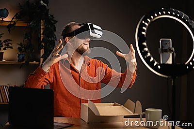 Handsome tech blogger male influencer unboxing device using modern smartphone while reviewing VR headset at home Stock Photo