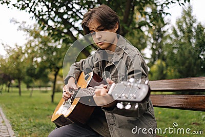 Handsome teenage playing acoustic guitar outdoor in Autumn time. Boy sitting on bench and playing music Stock Photo