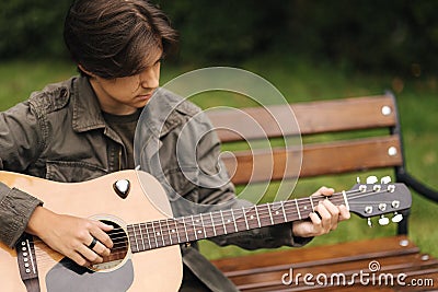Handsome teenage playing acoustic guitar outdoor in Autumn time. Boy sitting on bench and playing music Stock Photo
