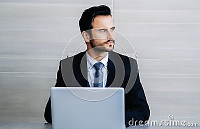 Handsome thoughtful bearded sitting at his desk while using laptop computer and looking away in modern office. People and Stock Photo