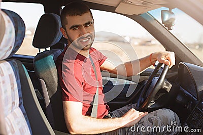 Handsome unshaven young male with stubble sits in car, has break after driving, looks at camera, enjoys high speed, being alone in Stock Photo