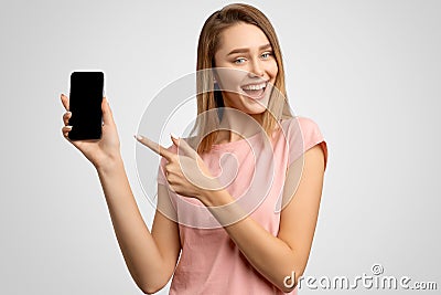 Handsome woman with index finger shows blank phone screen where you can place your ad. Positive young girl smiles snow-white smile Stock Photo