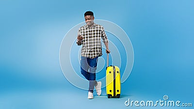 Handsome young black man with suitcase using mobile phone, booking summer vacation tourist trip over blue background Stock Photo