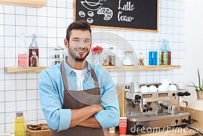 handsome young cafe owner in apron standing with crossed arms and smiling Stock Photo