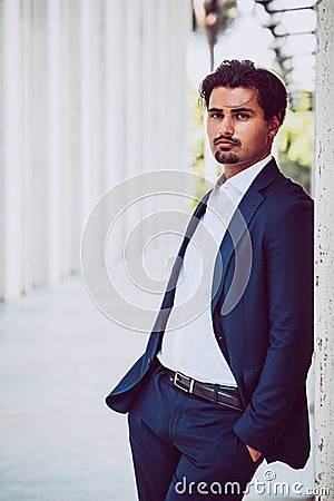 Handsome young elegant man posing outdoors along a corridor of white columns. Fashionable hair wearing jacket, pants and white Stock Photo