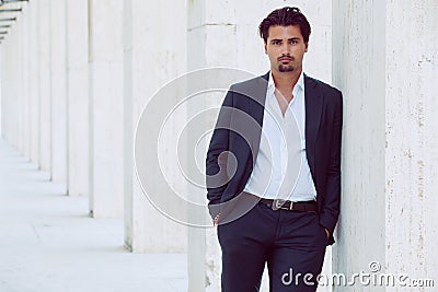 Handsome young elegant man posing outdoors leaning on a column along a corridor. Fashionable hair. Stock Photo