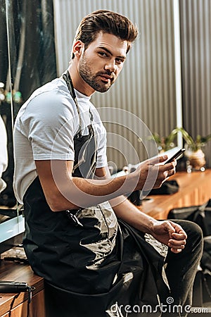 Handsome young hairstylist using smartphone and looking at camera Stock Photo