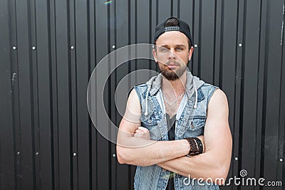 Handsome young hipster man in a fashionable blue denim vest in a t-shirt with a beard in a black stylish cap poses Stock Photo