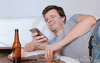 Handsome young man bachelor eating chips and drinking beer and browsing his mobile phone. Stock Photo