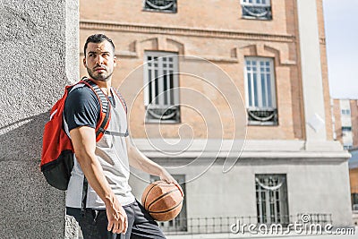 Handsome young man with a basketball and a backpack on his back looks aside while leaning on a column Stock Photo