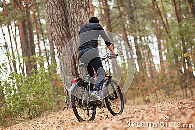 Handsome young man biking in countryside, guy wearing black sportwear and cap posing backwards around trees in wood, spending Stock Photo