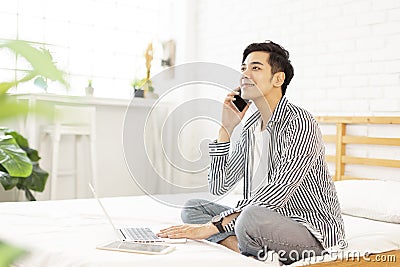 Handsome young man in casual clothes and working at home while sitting on the bed,with a laptop and talking on phone in bedroom Stock Photo