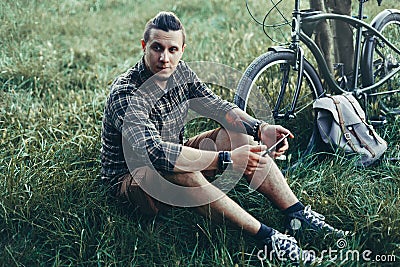 Handsome Young Man Cyclist Sits On Summer Meadow Near Bicycle, Holding At Tablet Recreation Resting Travel Destination Concept Stock Photo