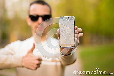 Handsome young man showing broken smartphone screen doing ok sign, thumb up with fingers Stock Photo