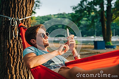 Handsome young man in sunglasses lying at sunset in a hammock and using a smartphone. The guy is resting in a hammock in the park Stock Photo