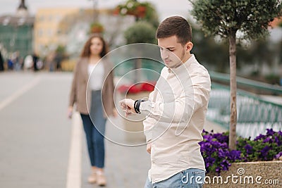 Handsome young man waiting for girlfriend outdoors and looking on the watch. Girl is coming to her boyfriend Stock Photo