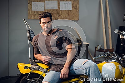 A handsome young motorcyclist in jeans and a t-shirt poses for a photo sitting on a bike in his garage Stock Photo