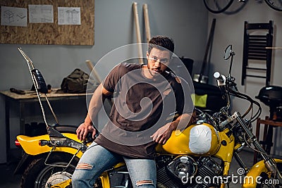 A handsome young motorcyclist in jeans and a t-shirt poses for a photo sitting on a bike in his garage Stock Photo