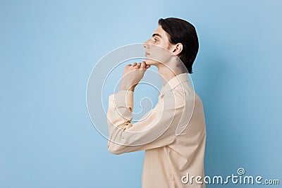 Handsome young thin dark-haired guy with blue eyes wearing yellow shirt holding hand on chin posing over blue wall. Side Stock Photo