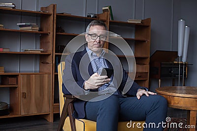 Hansome elderly businessman in blue suit and glasses working and looking on his mobile phone on office wall with boockshelf Stock Photo