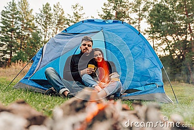 Happiness couple sitting at the entrance of the tent with cups in their hands. Travelers drink tea outdoors. Stock Photo