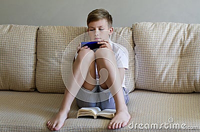 Teen boy is using phone with open book lying on the sofa. Stock Photo